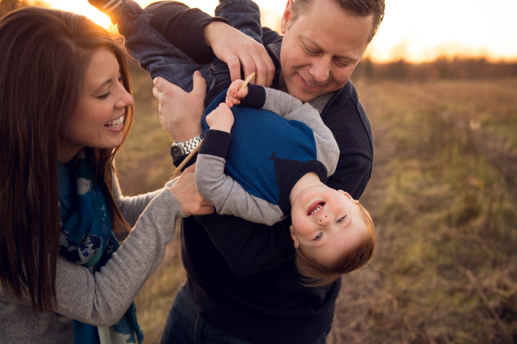 Smiling parents hold their laughing young child upside down in a sunlit grassy field